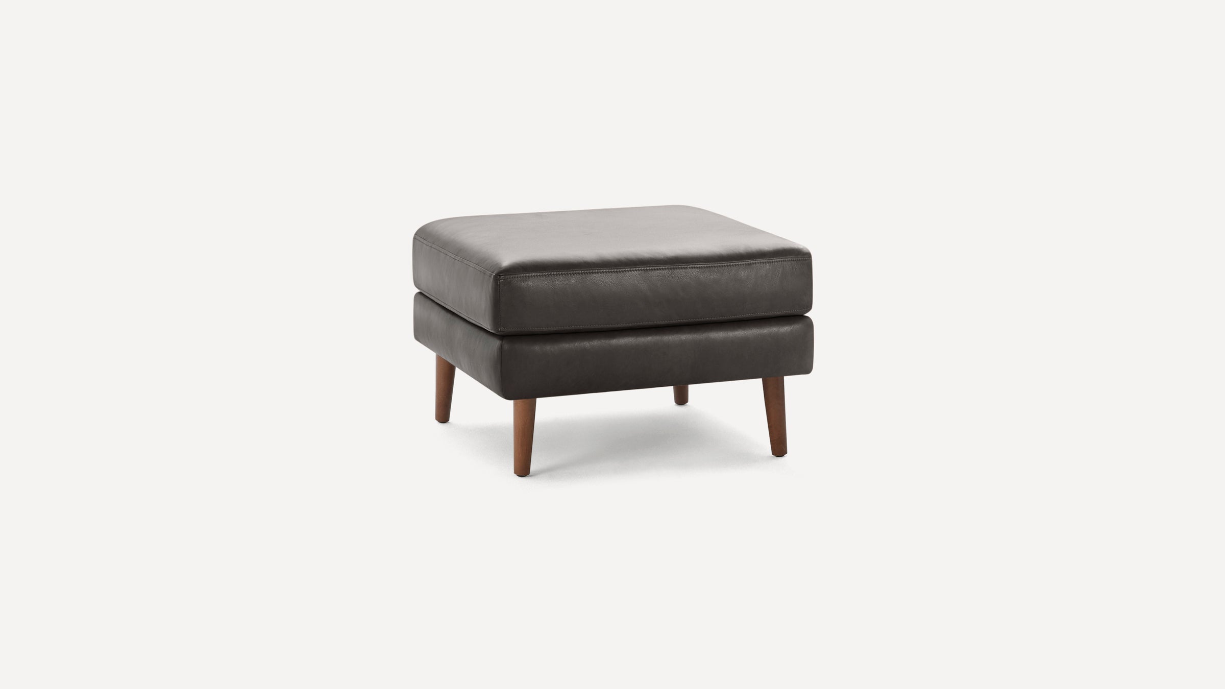 Square dark gray leather ottoman with four wooden legs against a white background.