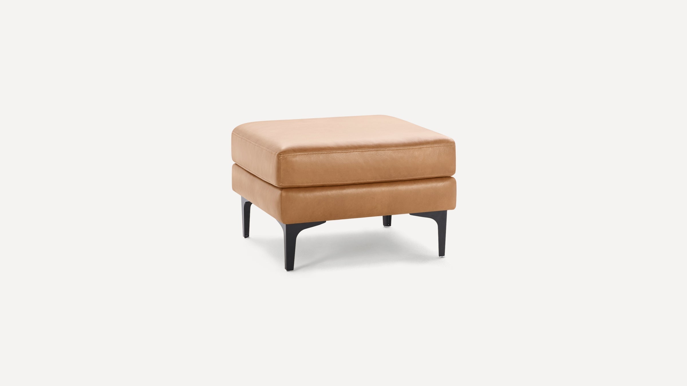 Square tan leather ottoman with cushioned top and black metal legs on a white background.