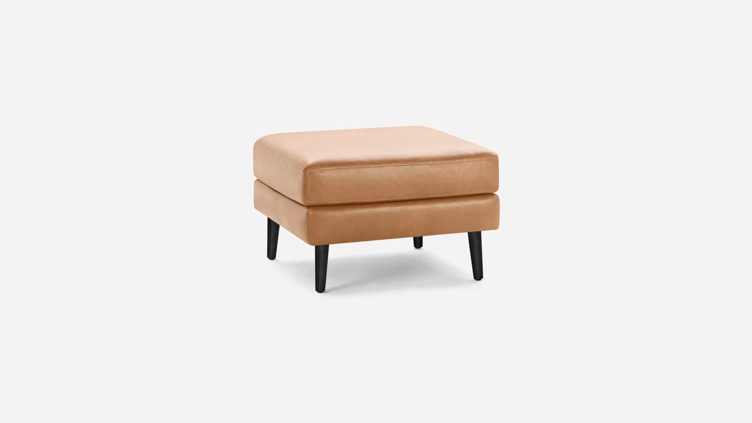 Square tan leather ottoman with cushioned top and four black tapered legs on white background.