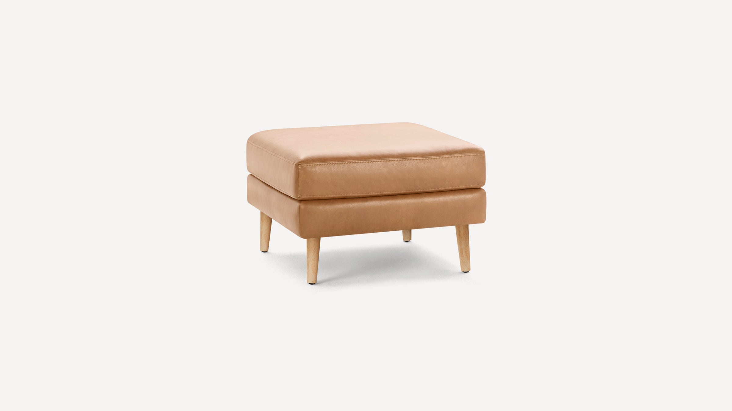 Square tan leather ottoman with cushioned top and four light wooden tapered legs on plain white background.