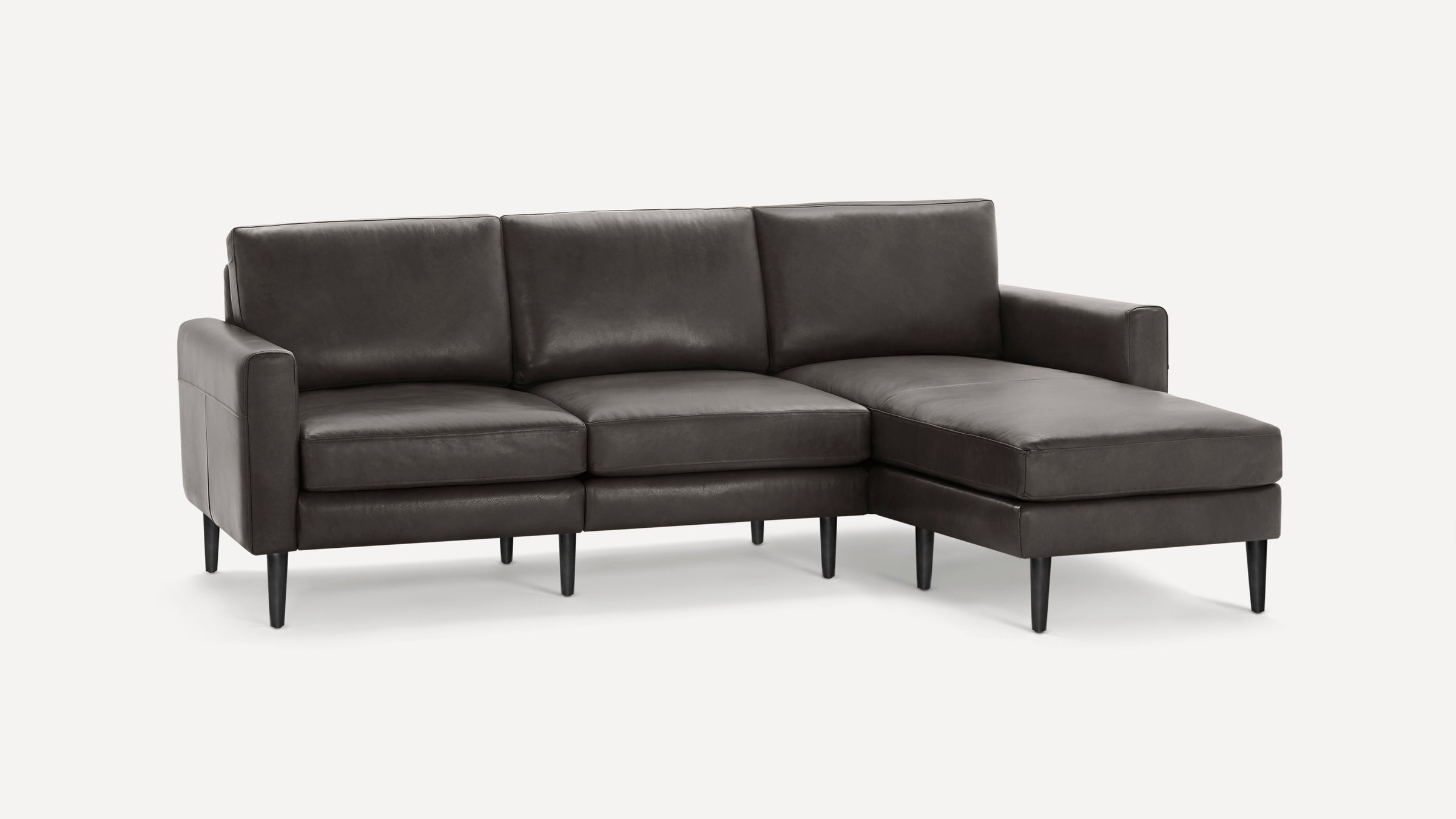 Modern dark gray leather sectional sofa with three seat cushions, chaise lounge on the right, and tapered black legs against white background.