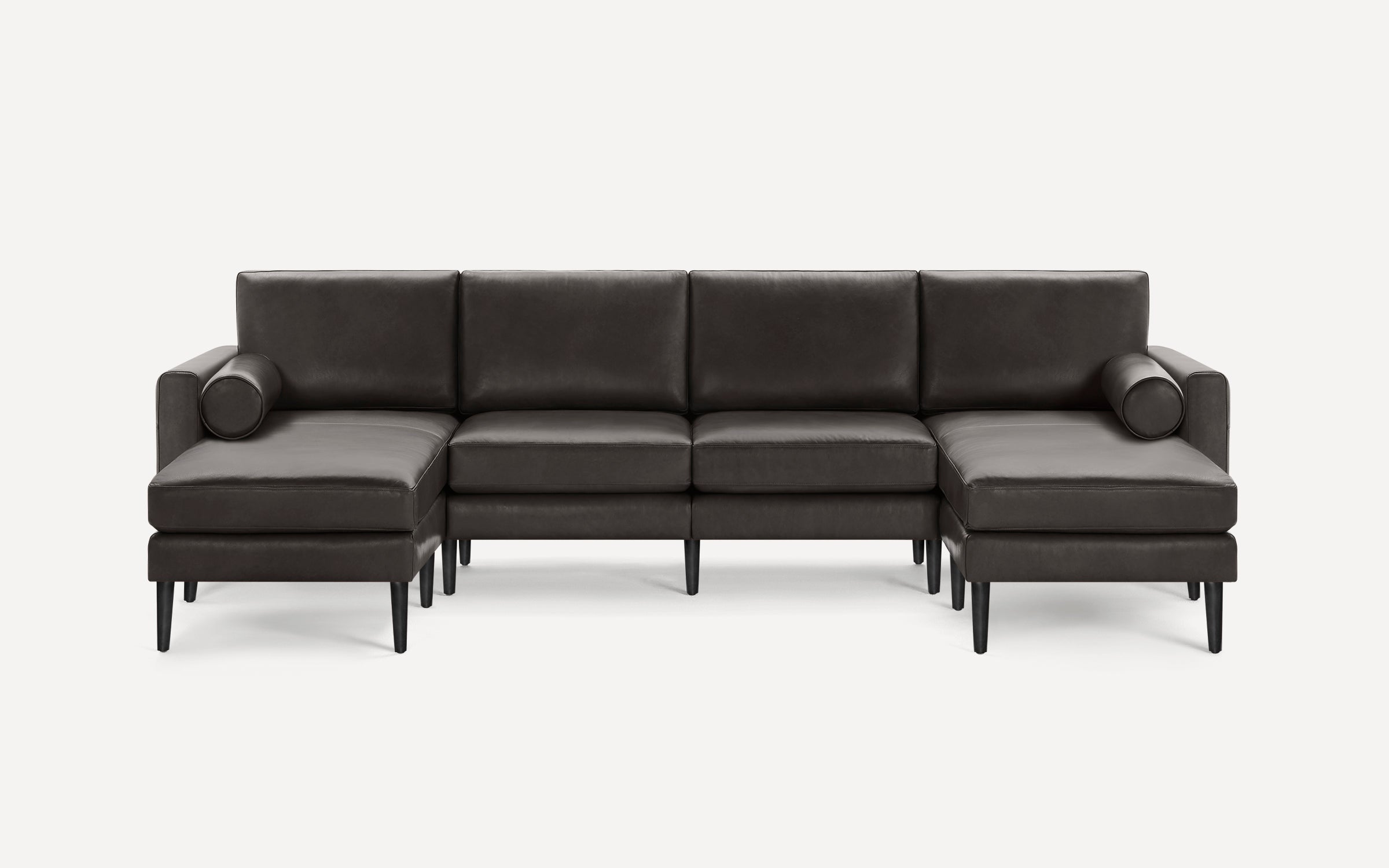 Modern black leather sectional sofa with chaise lounge ends and cylindrical bolster pillows, supported by slender black legs.