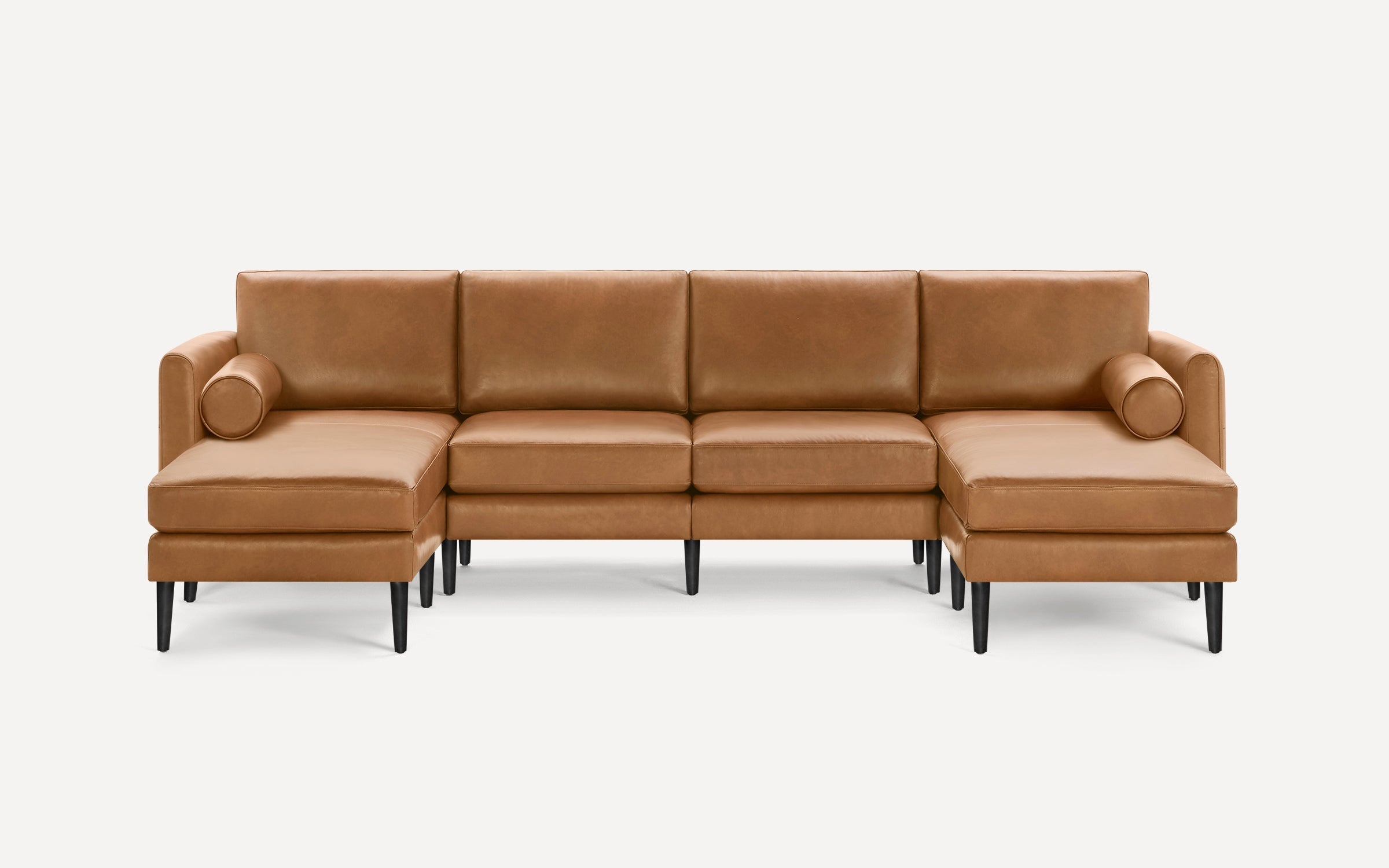 Modern sectional sofa in tan leather with clean lines, black tapered legs, and two cylindrical bolster pillows on each chaise end.