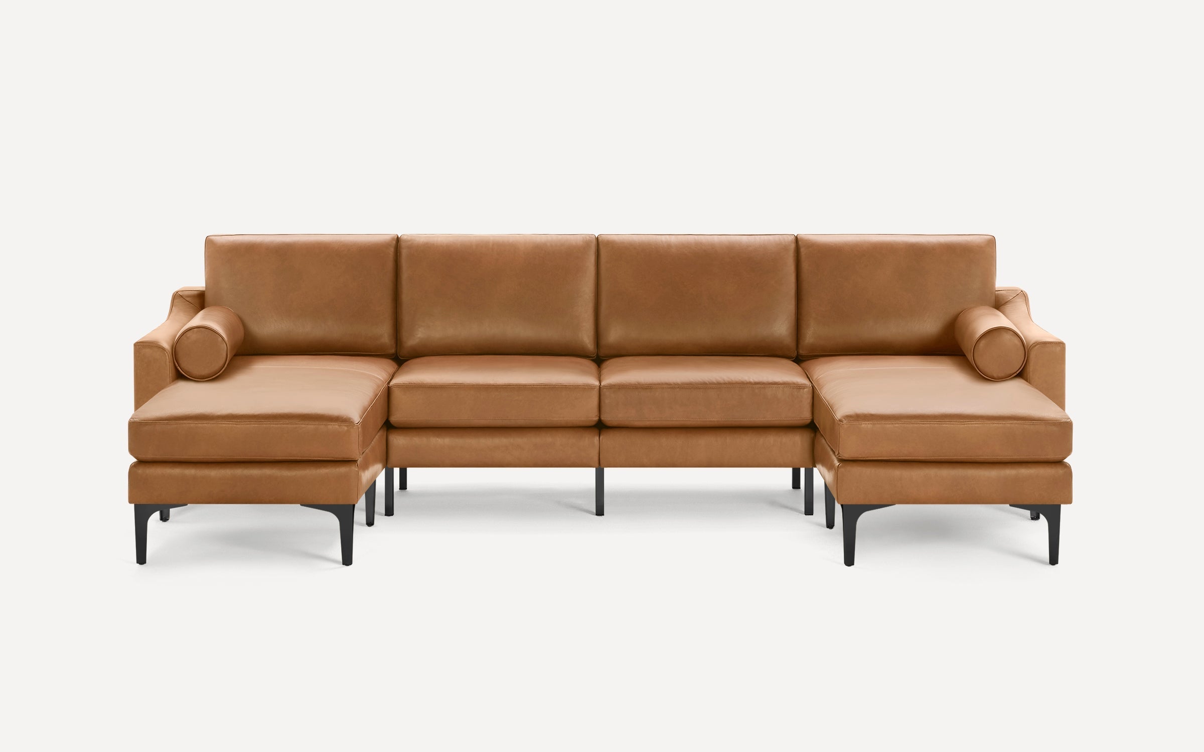 Modern sectional sofa in tan leather with clean lines, black metal legs, and two cylindrical bolster pillows on each end.