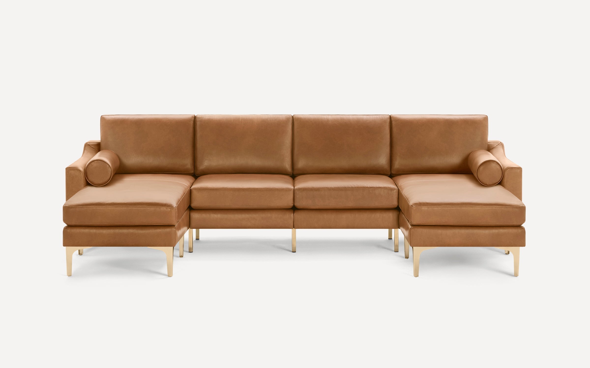 Tan leather U-shaped sectional sofa with gold metal legs and cylindrical bolster pillows on each end.