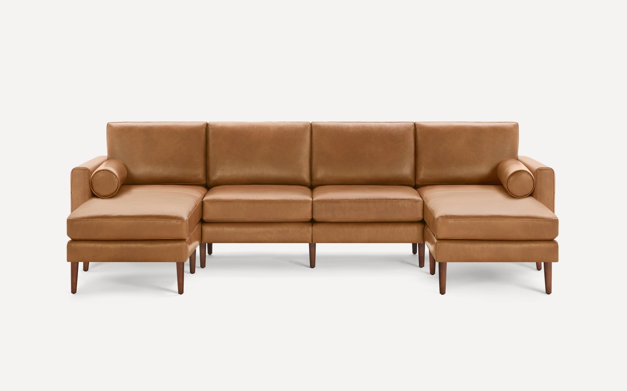 Modern mid-century tan leather sectional sofa with clean lines, wooden legs, and two cylindrical bolster pillows.