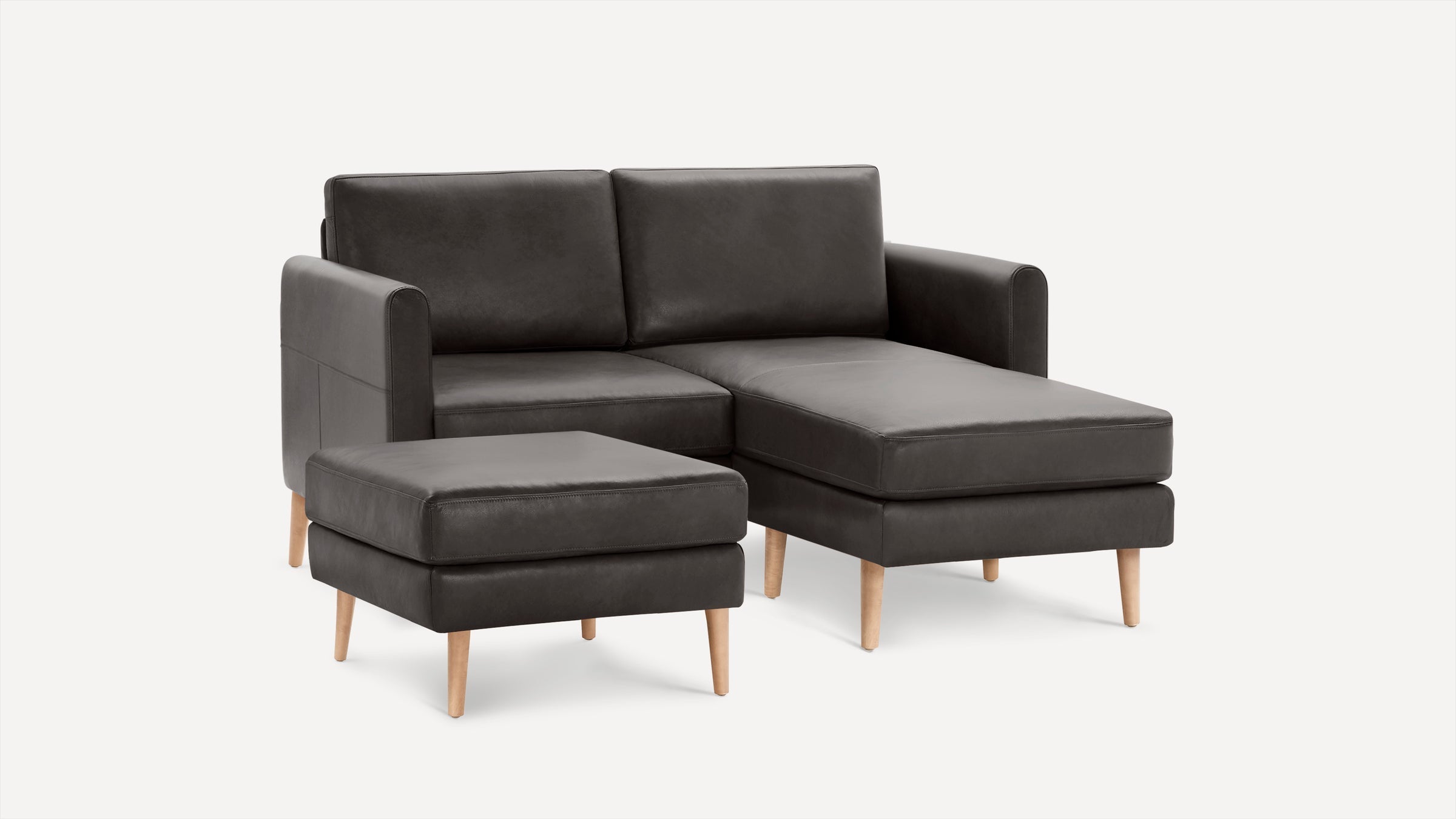 Dark gray leather sectional sofa with chaise lounge and matching ottoman, featuring light wood tapered legs on a white background.