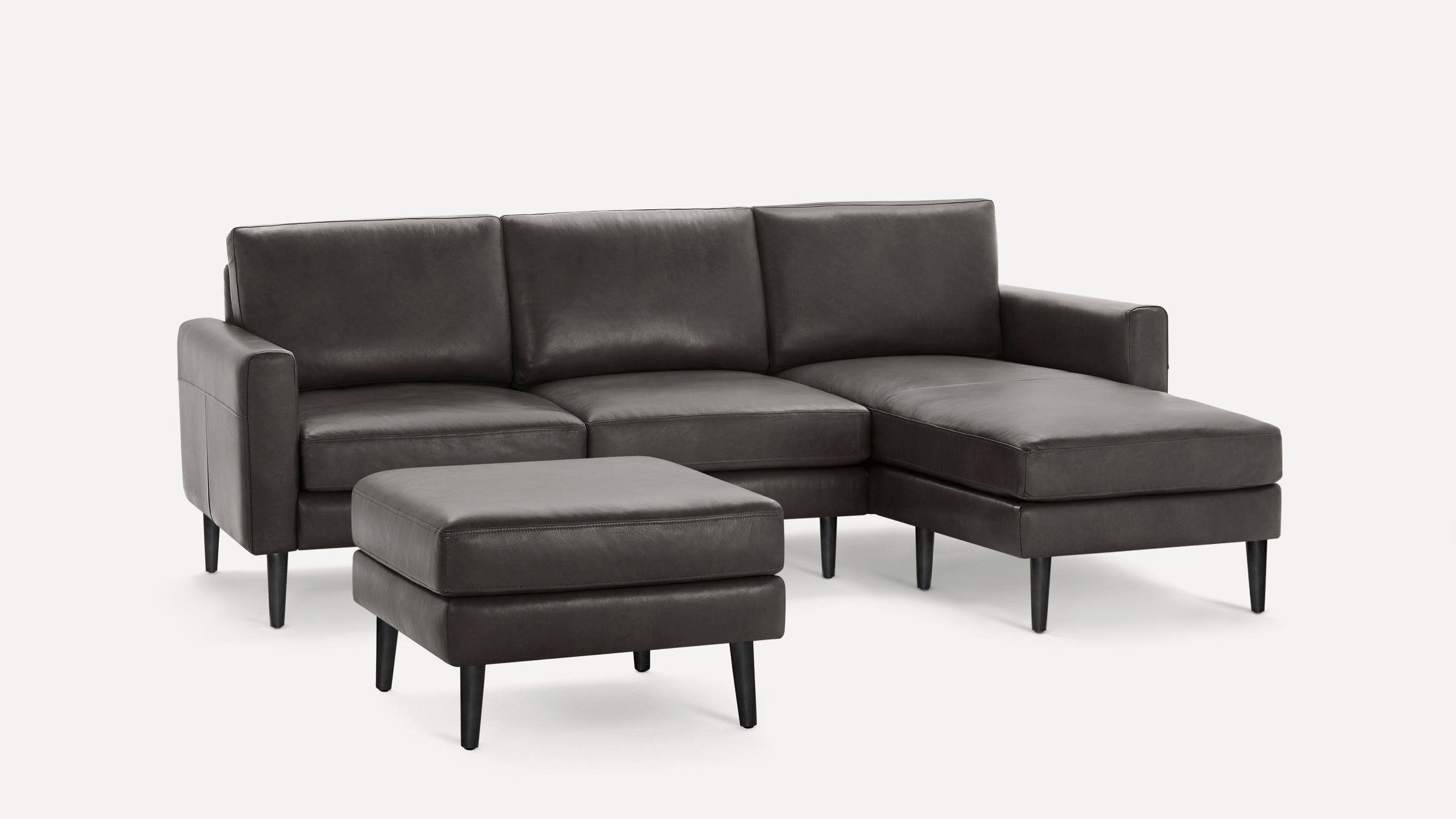 Dark gray leather sectional sofa with chaise lounge and matching ottoman, featuring slim black wooden legs.