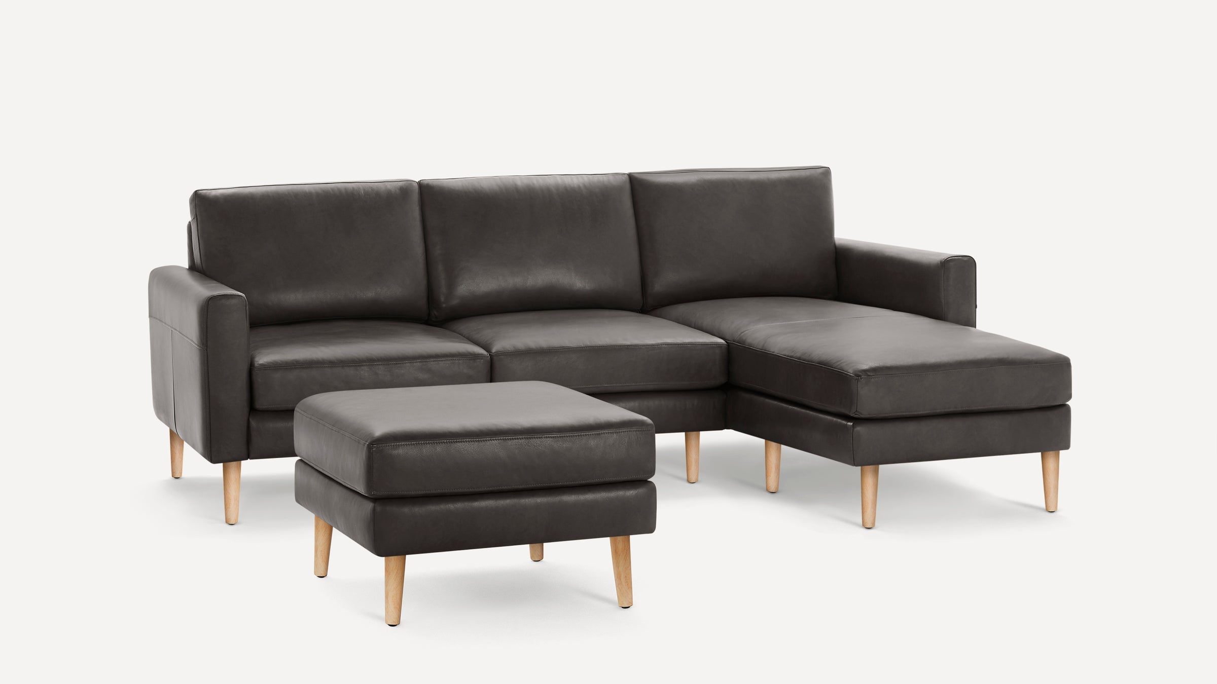 Dark gray contemporary leather sectional sofa with wooden legs and matching ottoman on white background.