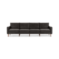 Modern long dark gray leather sofa with four seat cushions, straight armrests, and wooden legs, isolated on a white background.