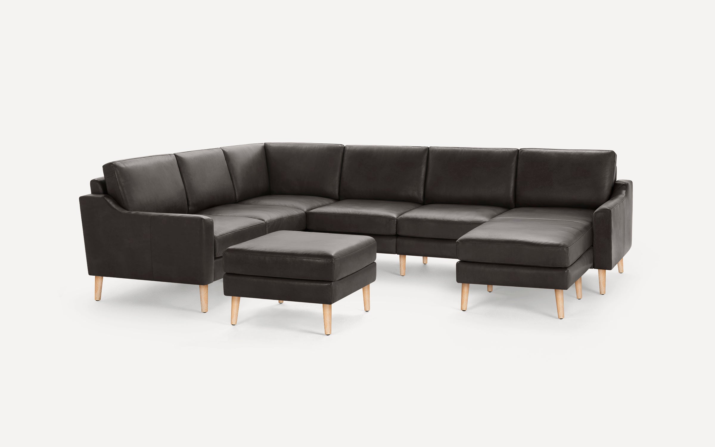 Dark gray leather sectional sofa with wooden legs, including a matching ottoman, on white background.