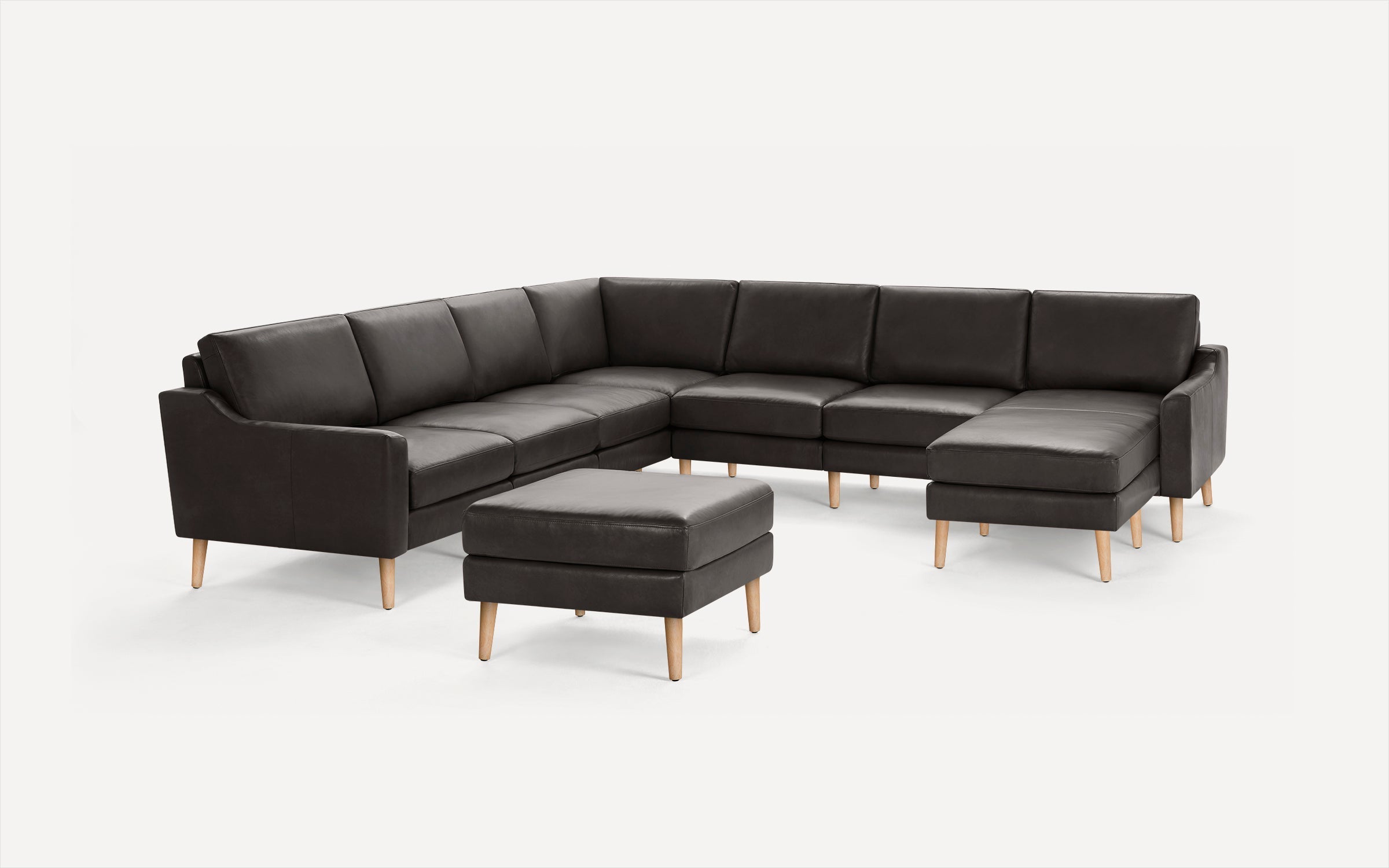 Dark gray leather sectional sofa with wooden legs and matching square ottoman on a white background.