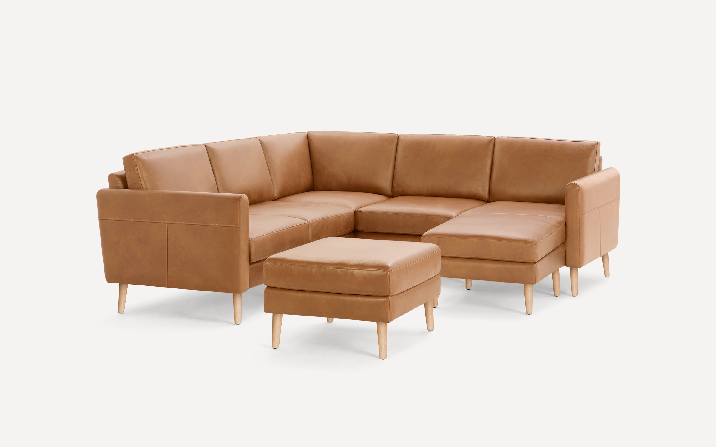 Light brown leather L-shaped sectional sofa with wooden legs and matching ottoman on white background.