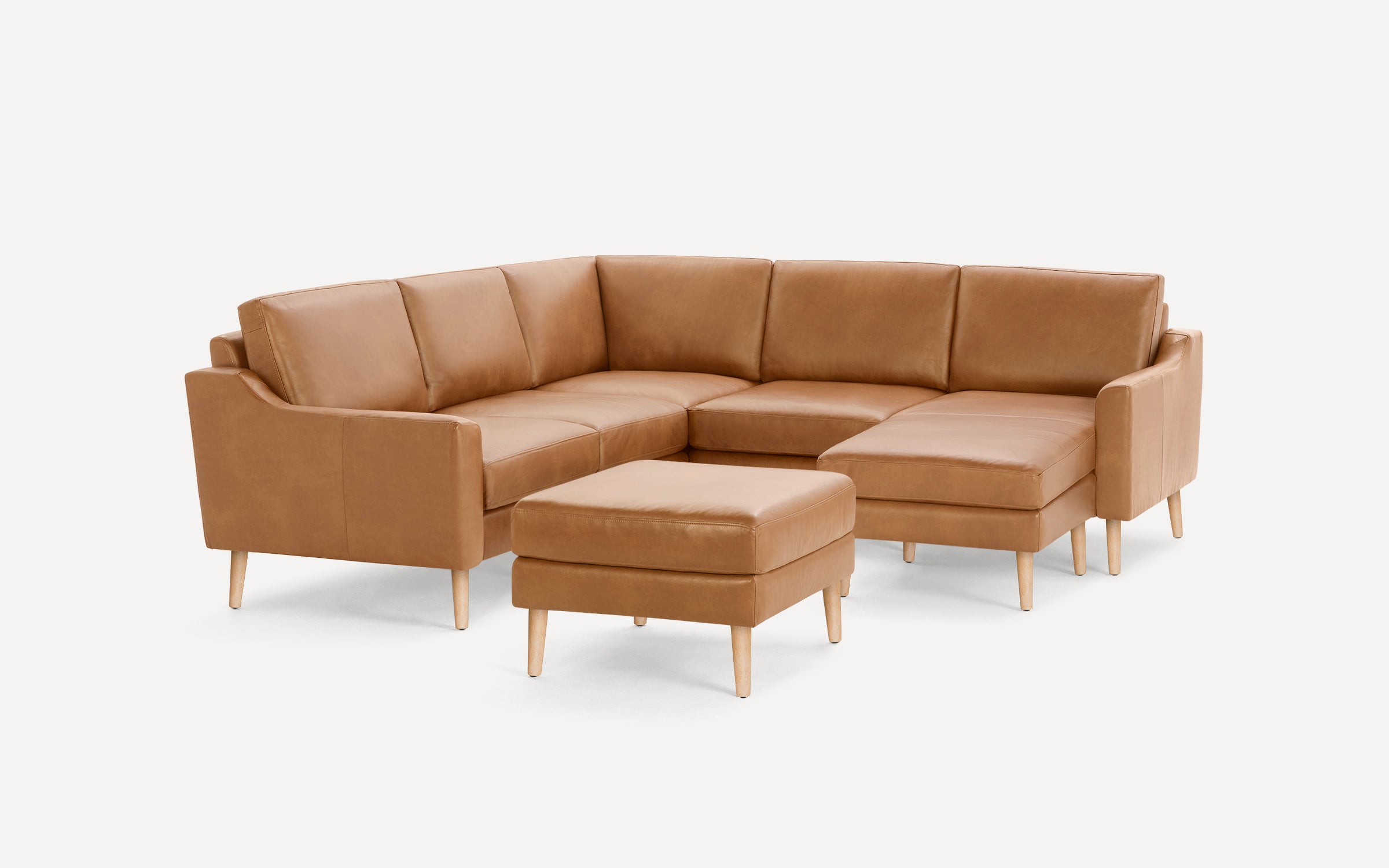 Modern tan leather sectional sofa with wooden legs and matching ottoman on a white background.