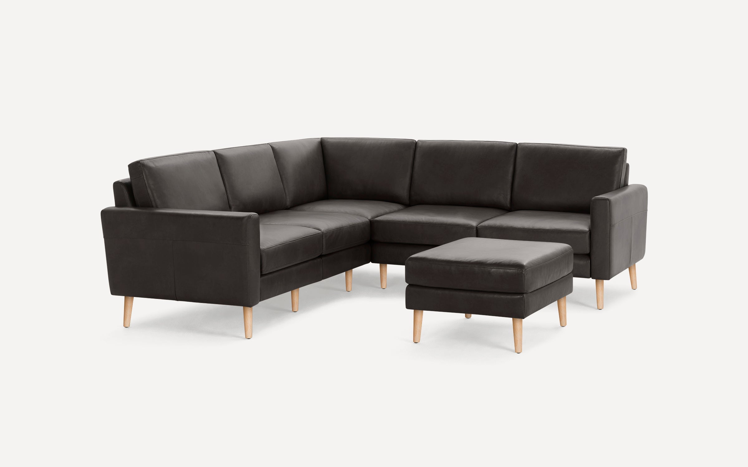 Dark gray leather sectional sofa with wooden legs and matching square ottoman on a plain white background.