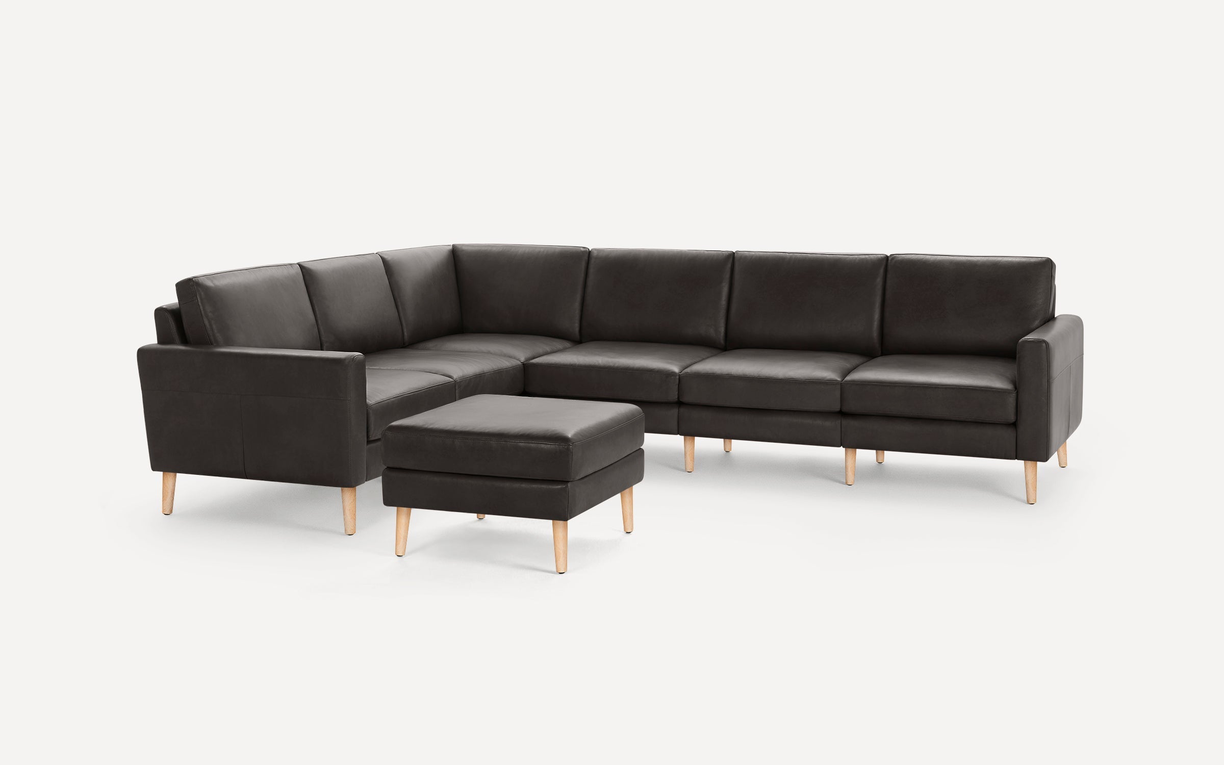 Black leather L-shaped sectional sofa with wooden legs and matching ottoman on plain white background.