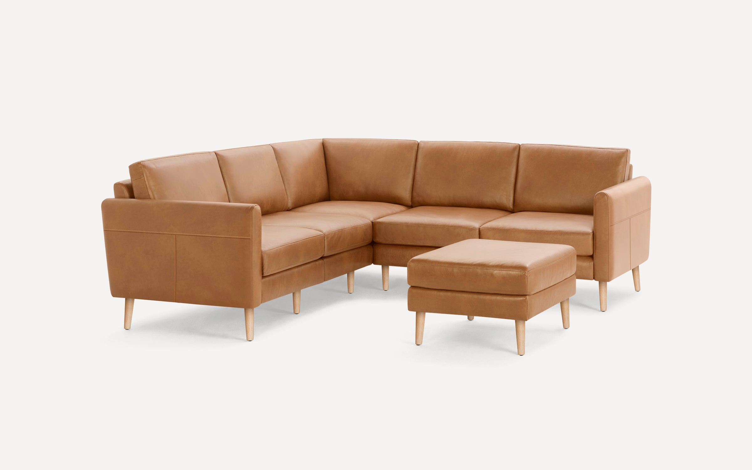 Modern camel brown leather sectional sofa with wooden legs and matching ottoman on white background.