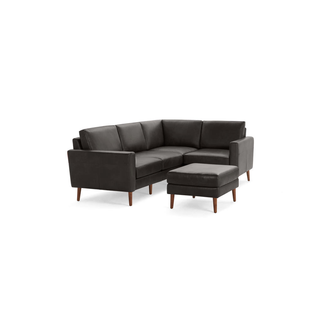 Dark gray leather L-shaped sectional sofa with wooden legs and matching ottoman against black background.