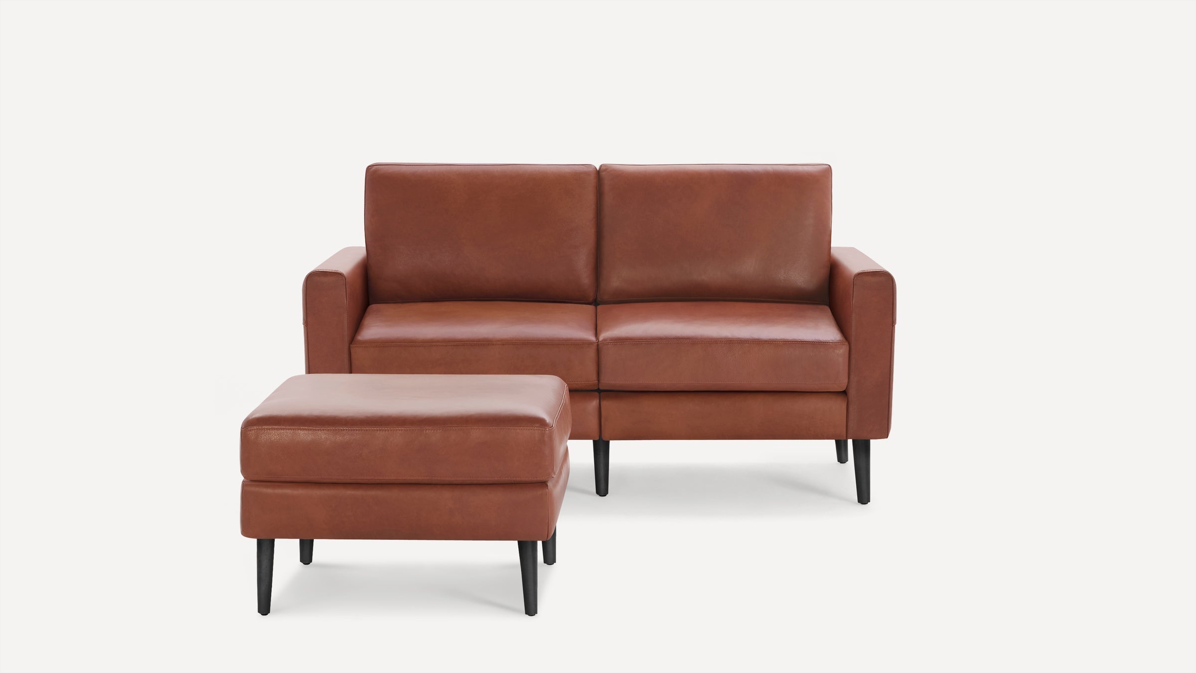 Brown leather loveseat with matching ottoman, both featuring black tapered wooden legs, set against a plain white background.