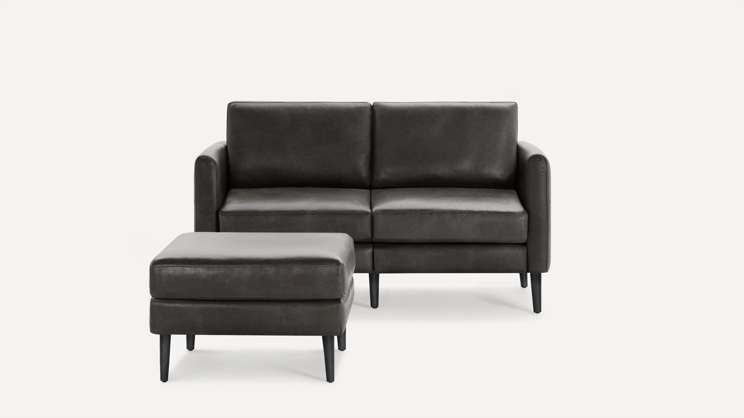 Modern dark gray leather loveseat sofa with matching cushioned ottoman, both featuring sleek black legs.