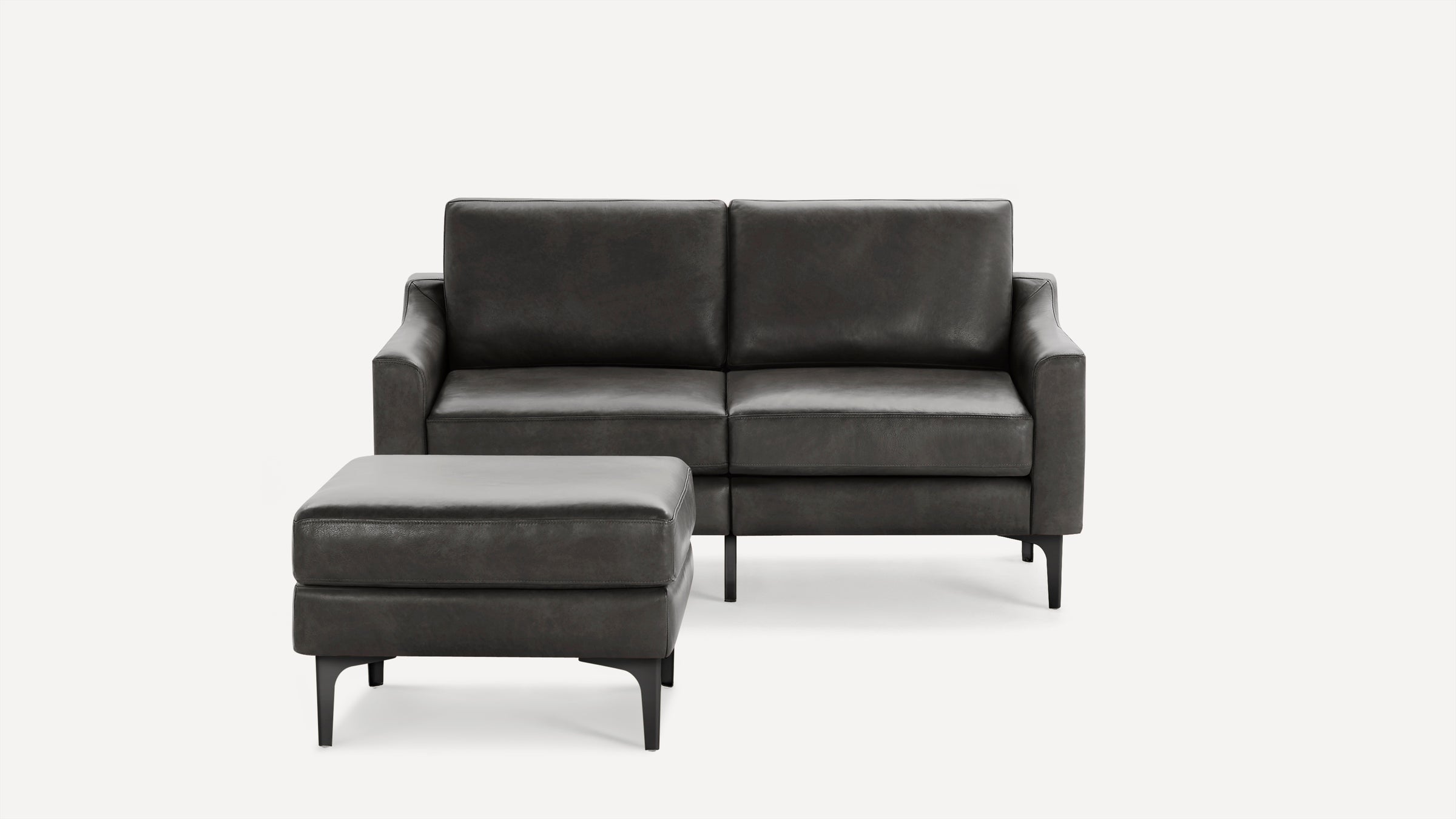 Modern dark gray leather loveseat with matching ottoman, featuring clean lines and black metal legs on a white background.