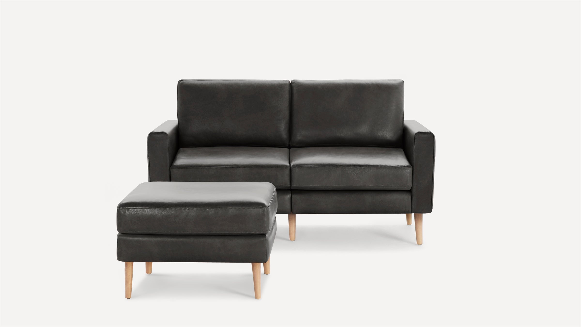 Dark gray leather two-seat sofa with matching ottoman, both featuring light wooden legs and clean, modern design.