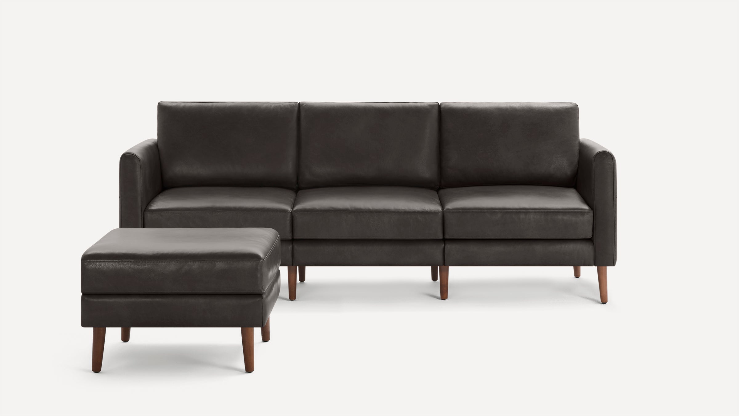 Dark gray leather three-seat sofa with armrests and matching ottoman, supported by wooden legs, on white background.