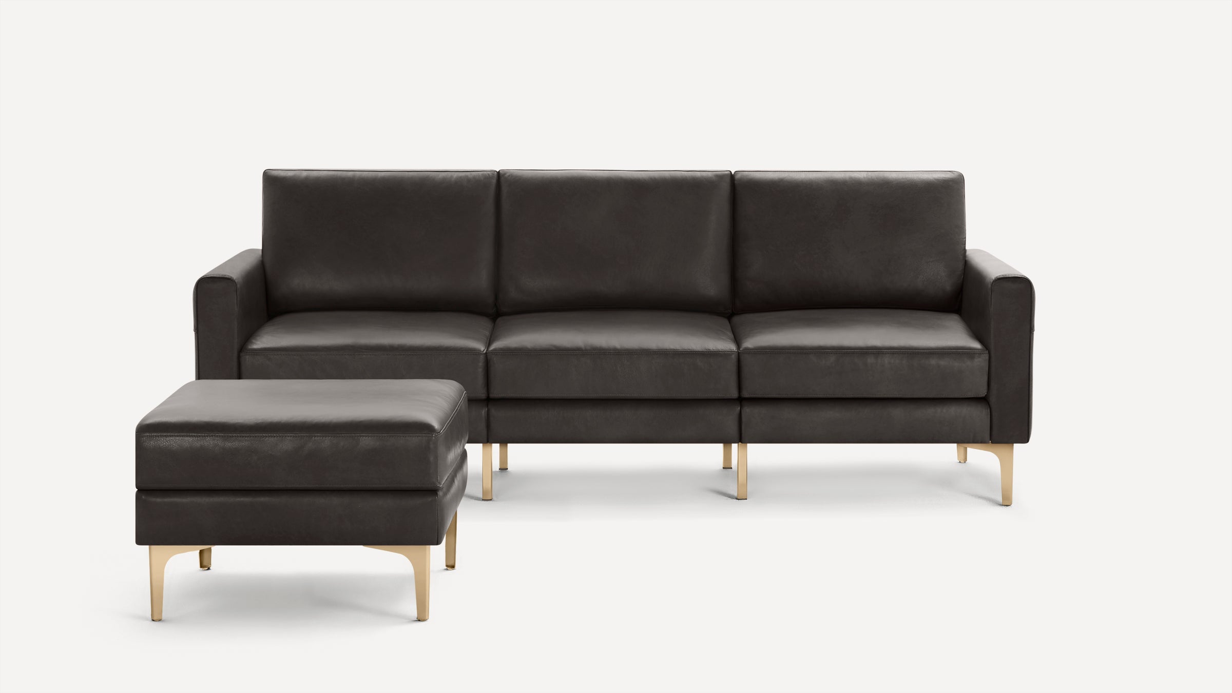 Dark gray leather three-seater sofa with matching ottoman, supported by sleek gold-colored legs, on a plain white background.