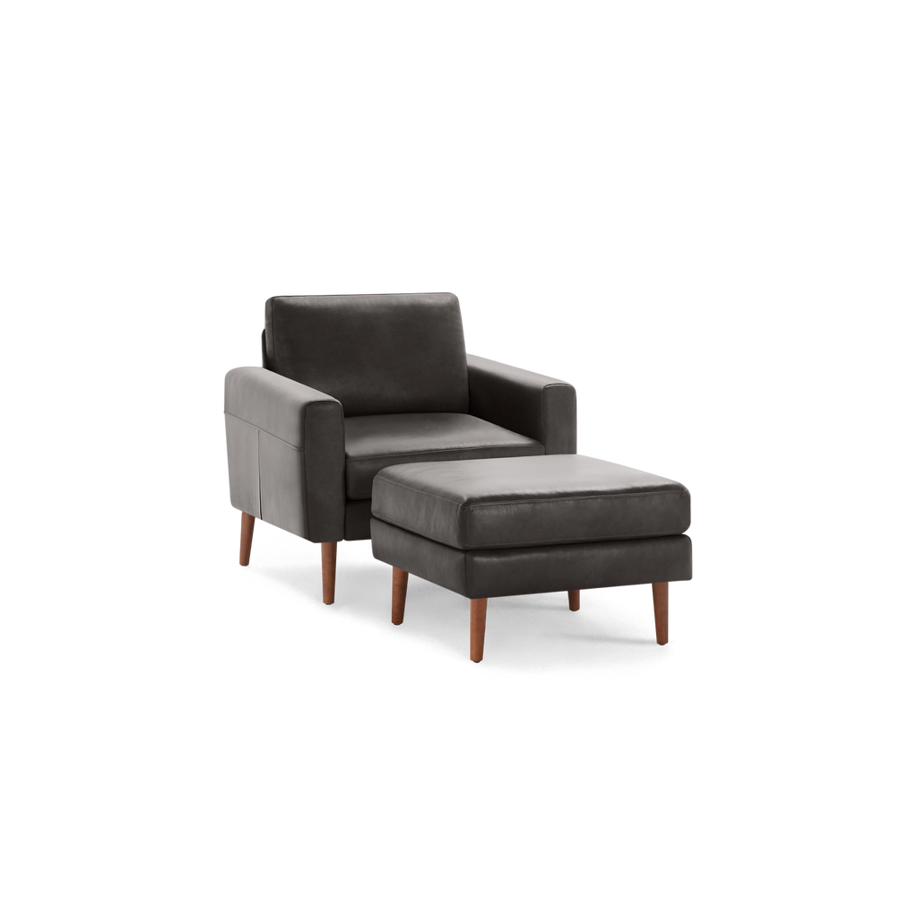 Dark gray upholstered armchair with matching ottoman, both featuring wooden tapered legs, pictured on a transparent background.