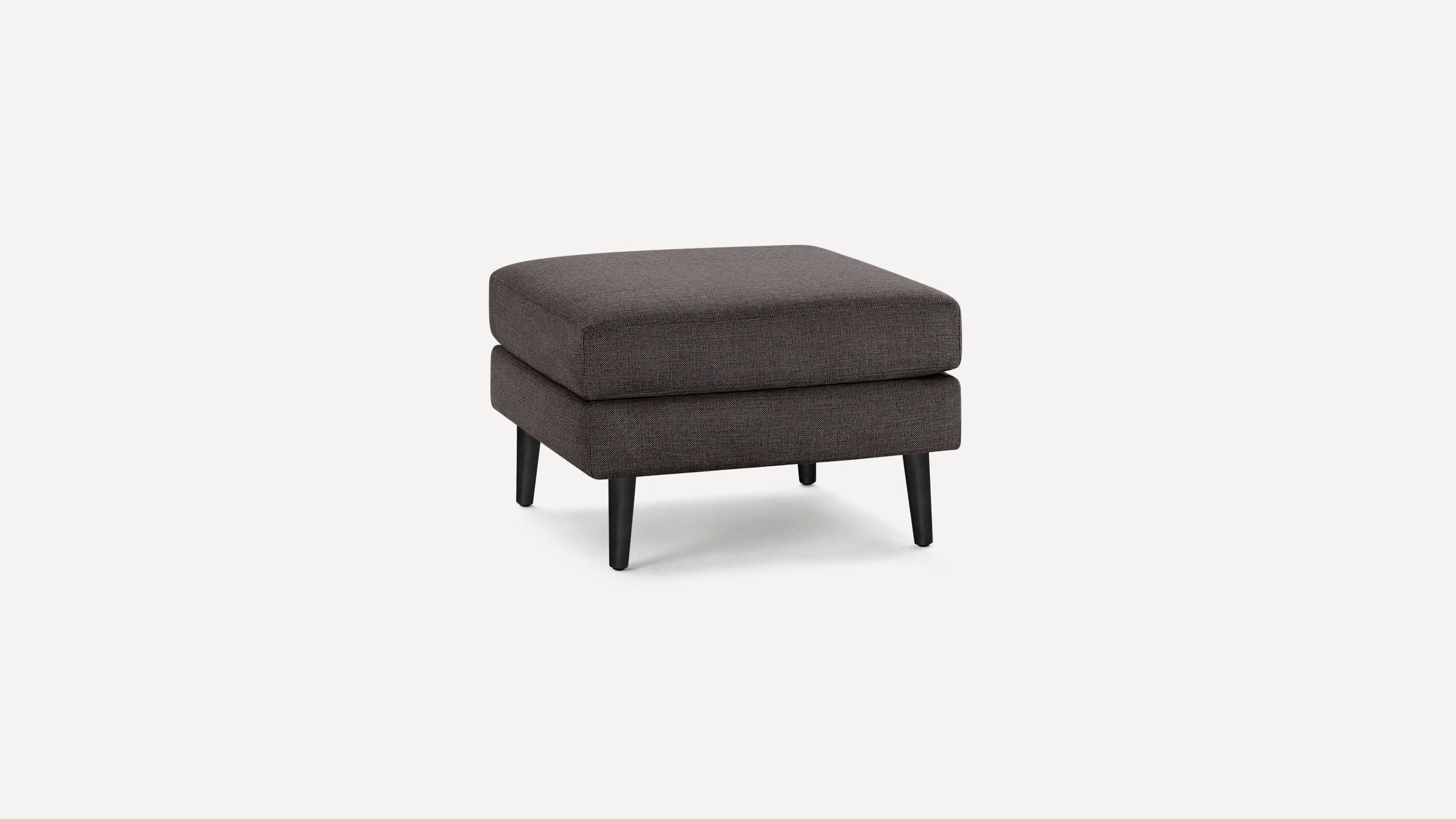 Dark gray upholstered square ottoman with thick cushion and four black tapered wooden legs on white background.