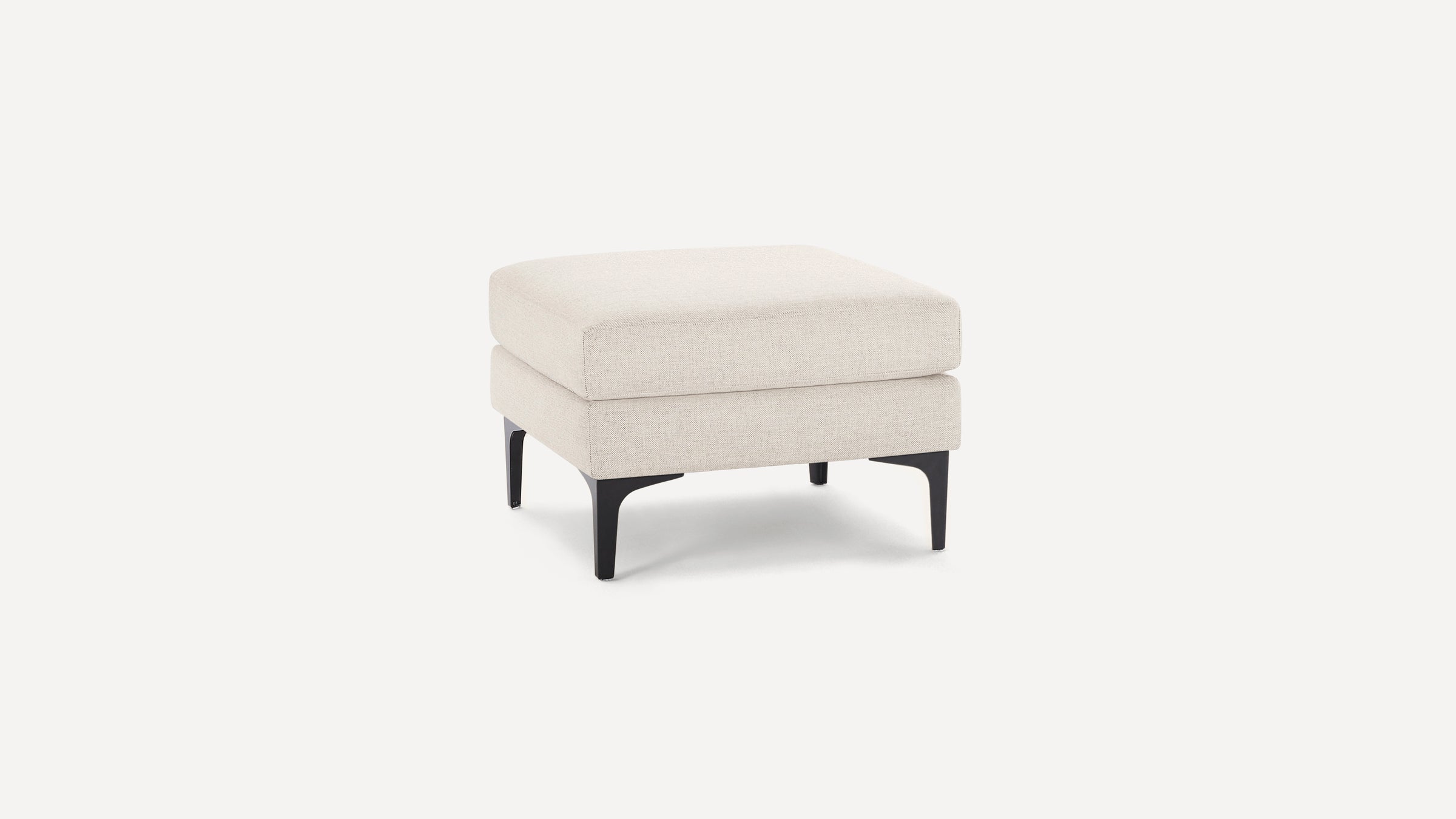 Square beige upholstered ottoman with cushioned top and black tapered legs on white background.