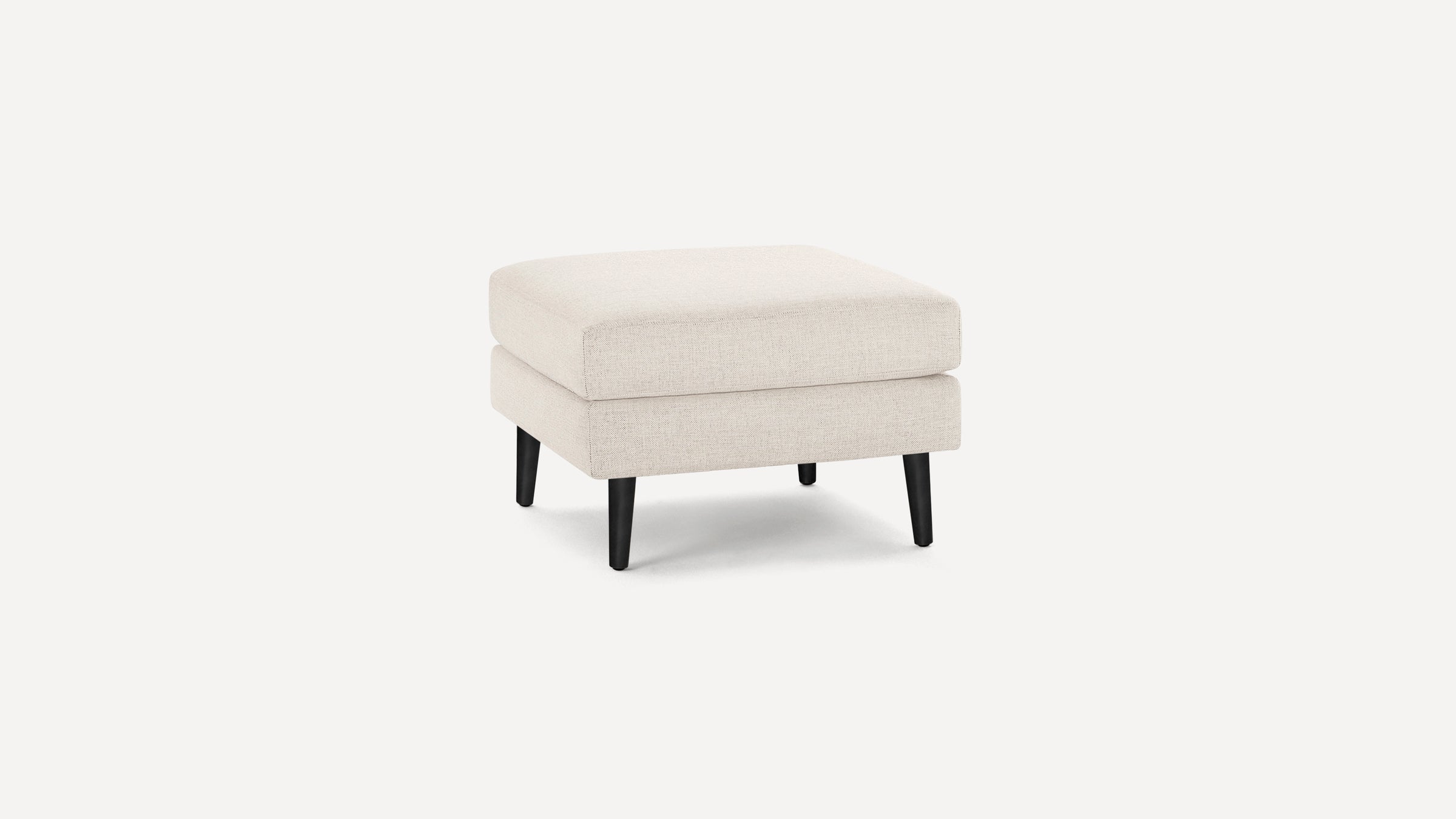 Square beige fabric ottoman with soft cushioning and four tapered black wooden legs on a white background.