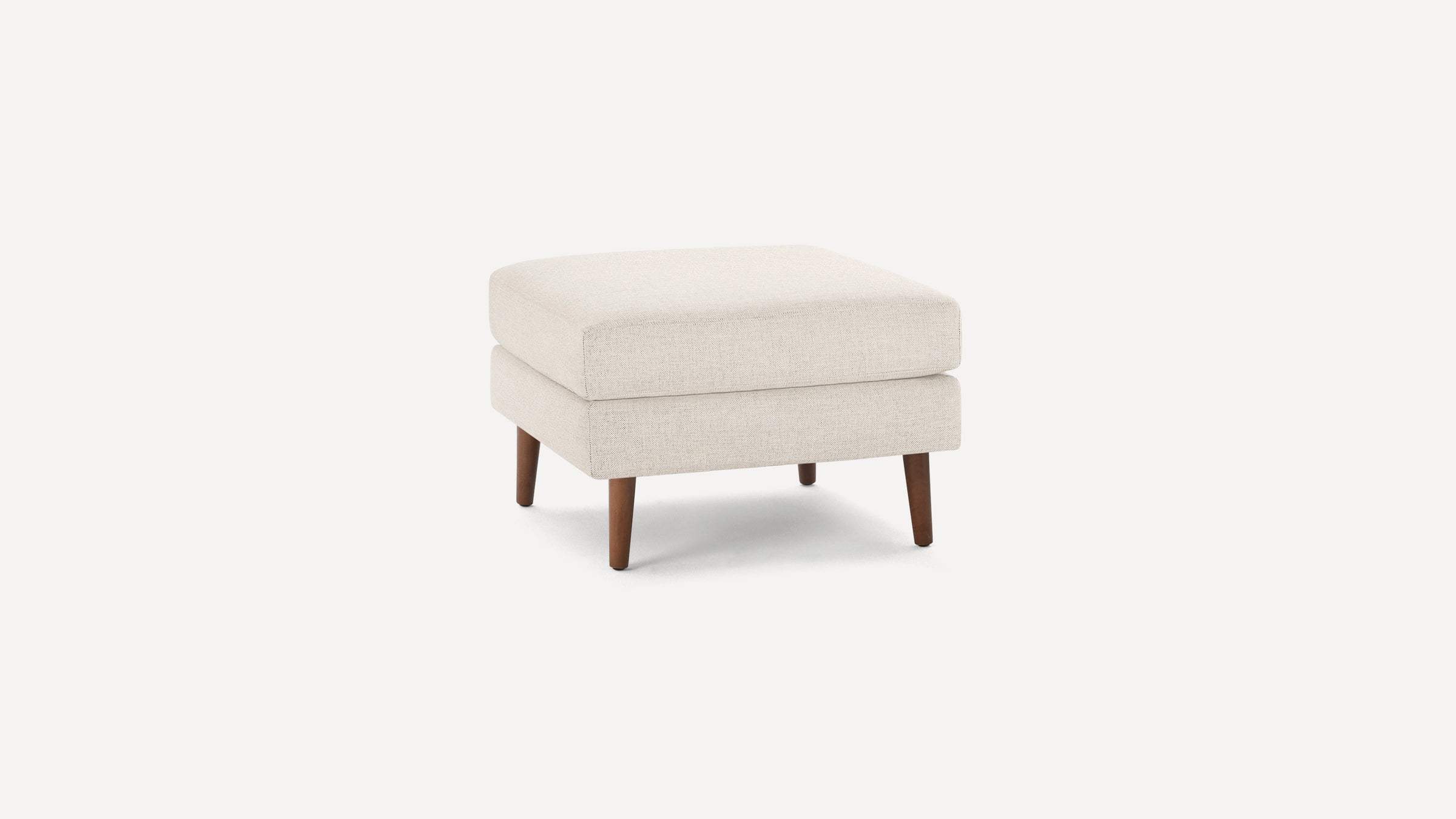 Square beige upholstered ottoman with wooden tapered legs, featuring a thick cushioned top for seating or resting feet.