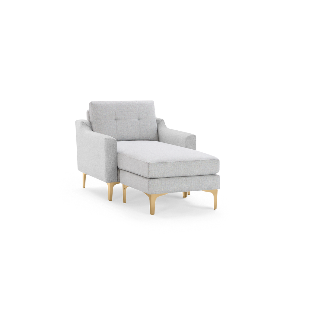 Light gray upholstered chaise lounge chair with tufted backrest and gold metal legs, shown on a black background.