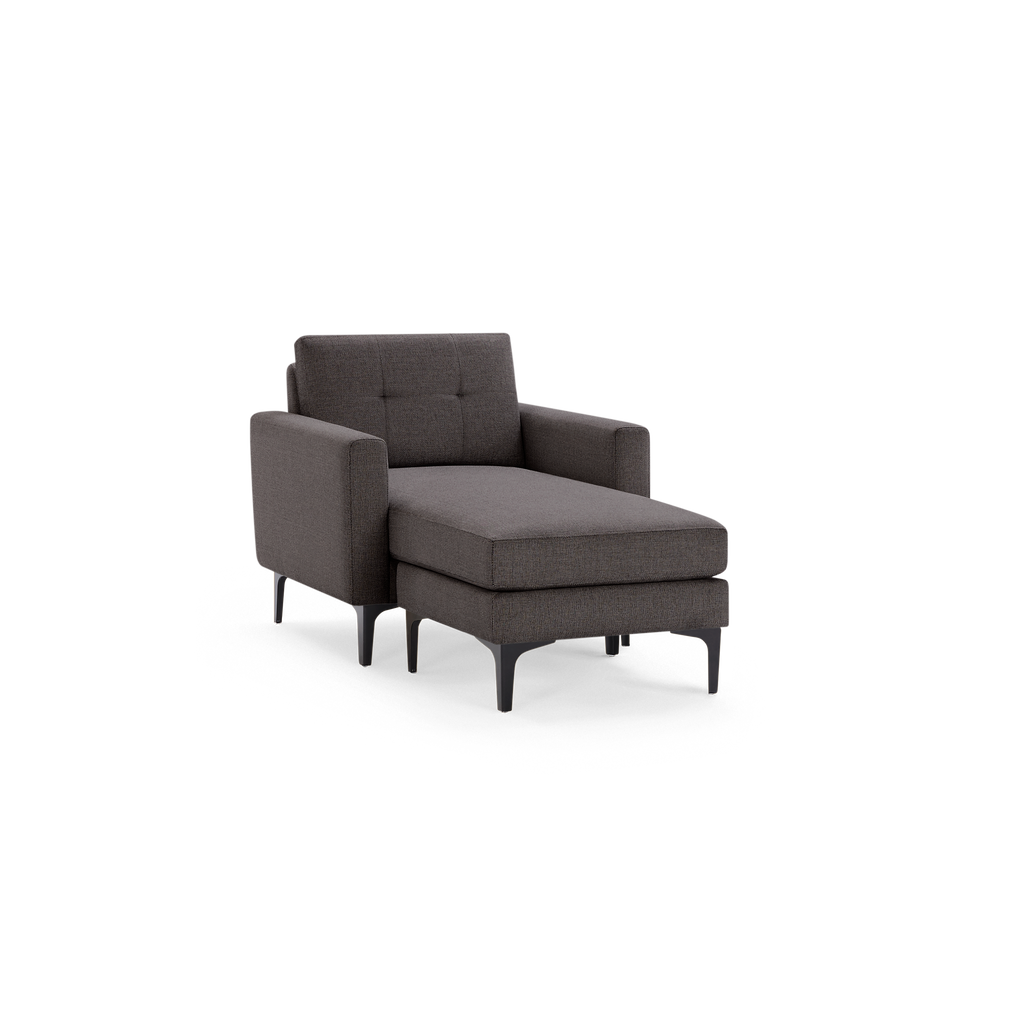 Dark gray upholstered chaise lounge chair with tufted backrest, high armrests, and black tapered legs on a black background.