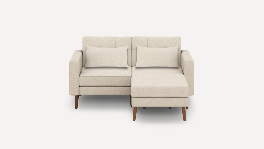 Beige two-seater sectional sofa with wooden legs, rectangular back cushions, and a chaise lounge on the right side against a plain background.