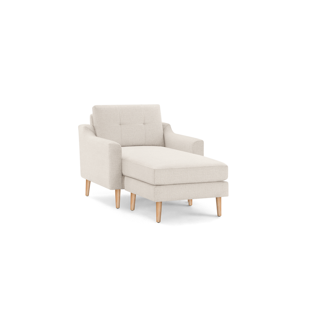 Beige upholstered chaise lounge chair with tufted backrest and light wooden legs on a black background.