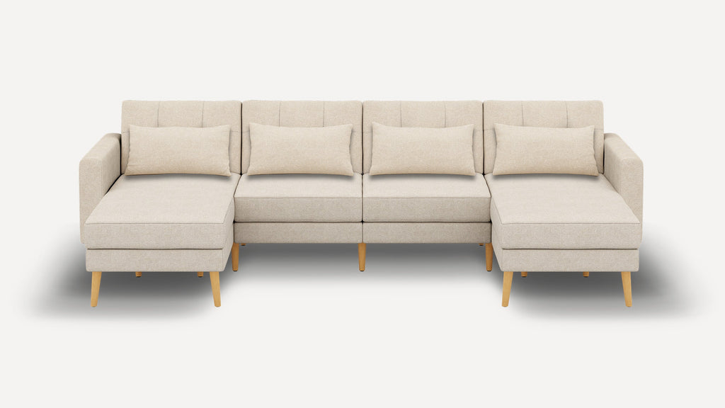 Sectional beige fabric sofa with wooden legs, featuring two chaise lounges and four matching lumbar pillows, set against a plain background.