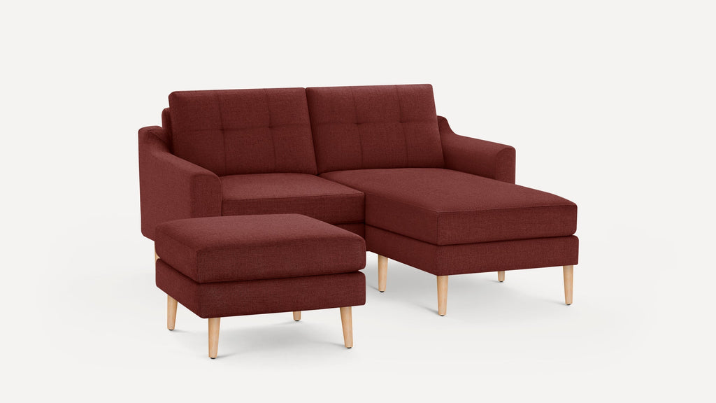Modern burgundy fabric sofa with chaise lounge and separate ottoman, featuring wooden tapered legs and tufted back cushions.