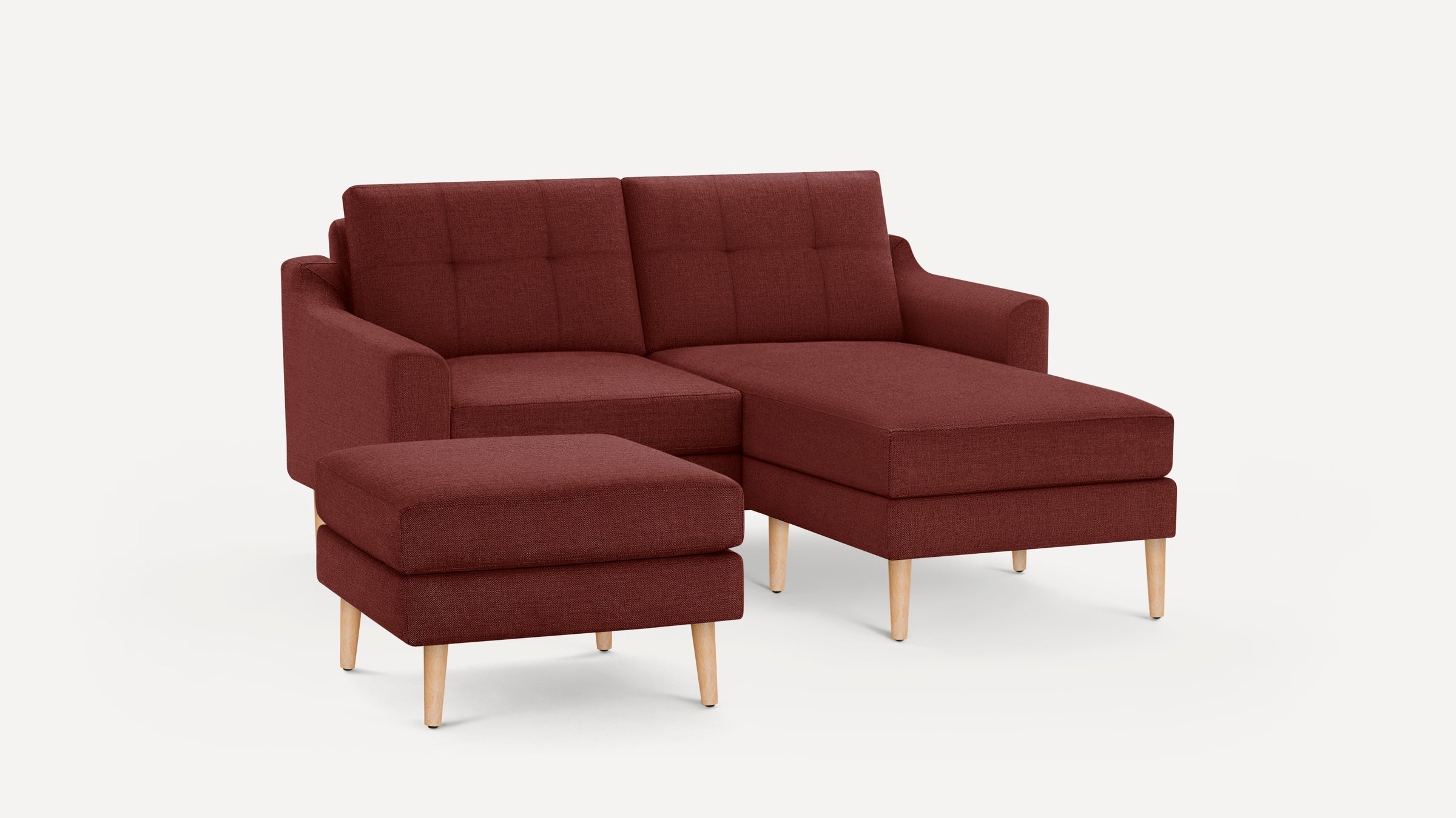 Modern burgundy fabric sofa with chaise lounge and separate ottoman, featuring wooden tapered legs and tufted back cushions.
