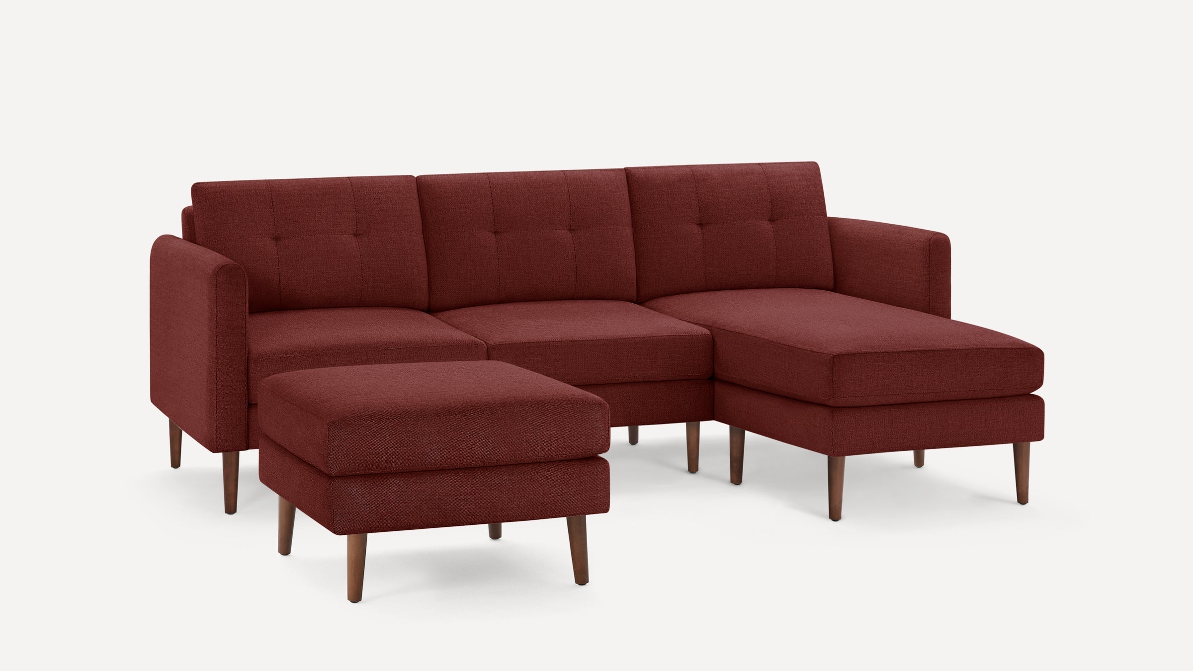 Modern L-shaped sectional sofa with tufted back cushions, deep red fabric upholstery, and wooden tapered legs, including a matching ottoman.