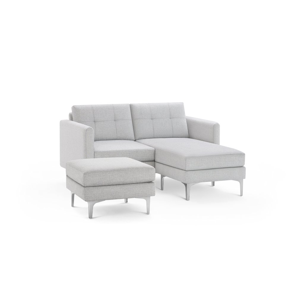 Light grey modern sectional sofa with tufted back cushions, chaise lounge on right side, and matching ottoman with metal legs.