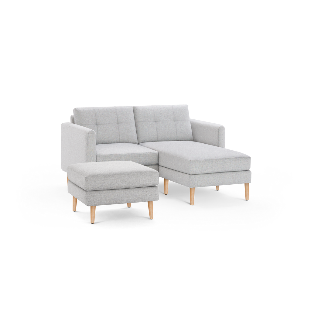 Light gray upholstered sectional sofa with tufted back cushions, wooden legs, and matching ottoman, featuring a right-side chaise lounge.