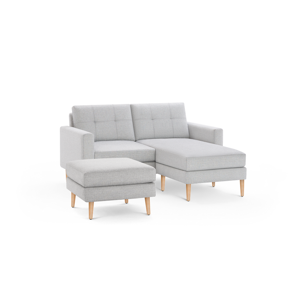 Light gray fabric chaise sofa with tufted back cushions, wooden legs, and a matching ottoman on a black background.
