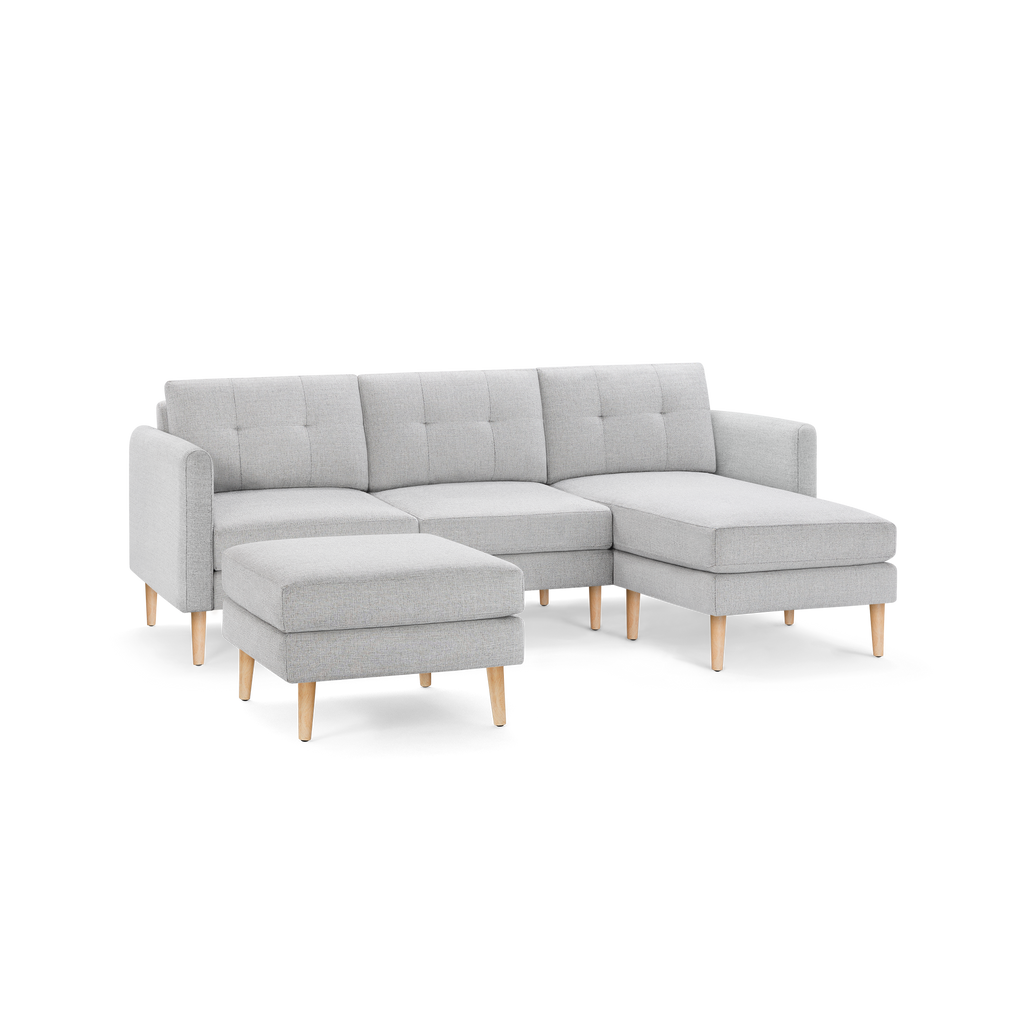 Light gray fabric L-shaped sectional sofa with tufted back cushions, matching ottoman, and wooden legs.