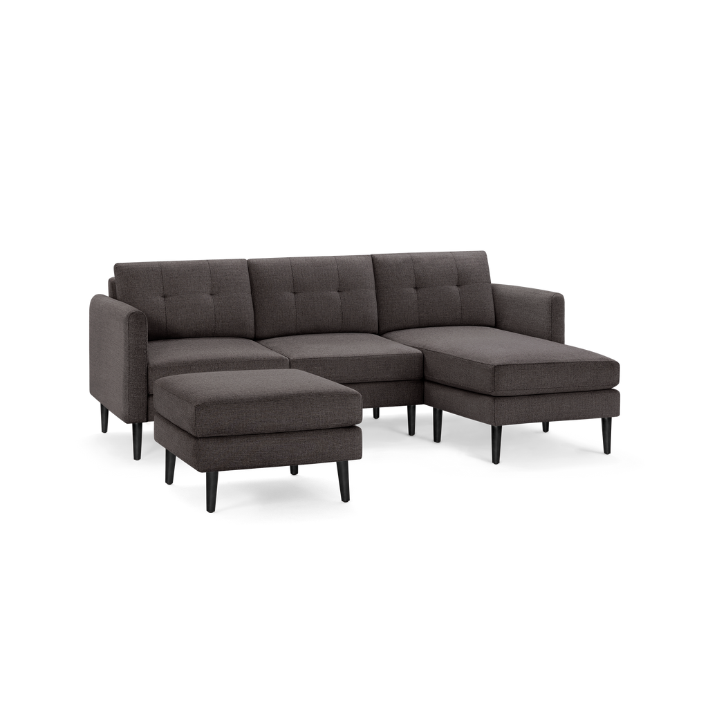 Dark gray modern sectional sofa with tufted back cushions, wooden legs, and matching ottoman on isolated background.
