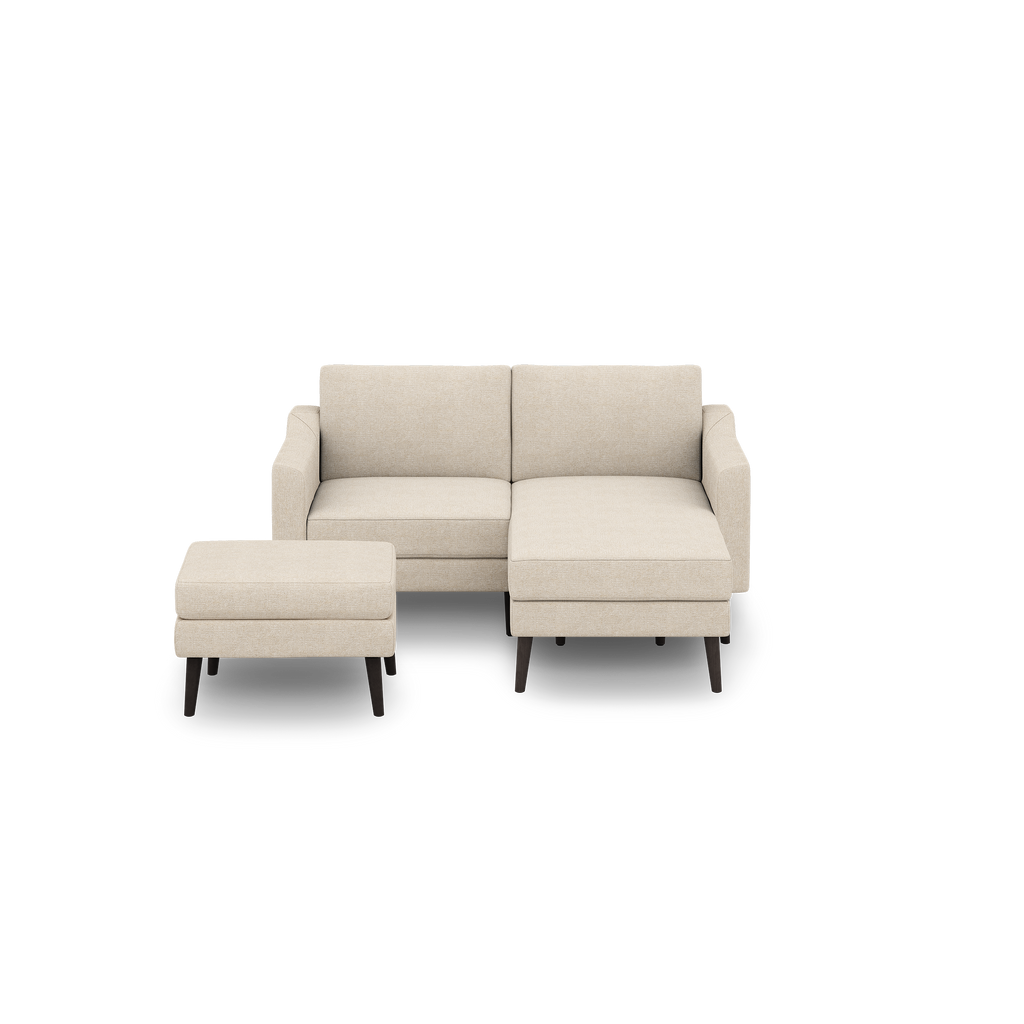 Beige fabric sectional sofa with chaise and matching ottoman, supported by dark wooden legs, against black background.