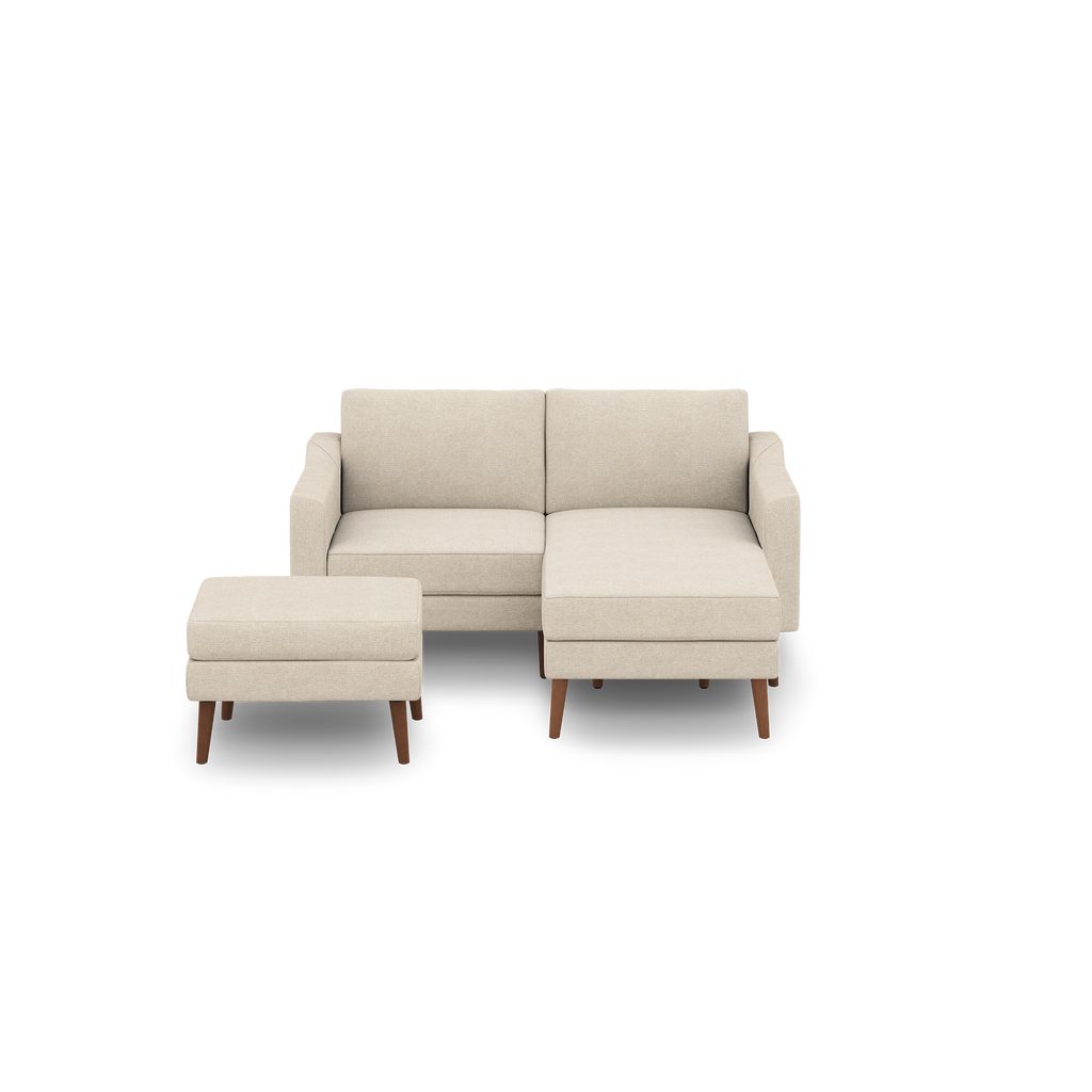 Beige fabric sectional sofa with wooden legs and matching ottoman, modern minimalist design, isolated on black background.
