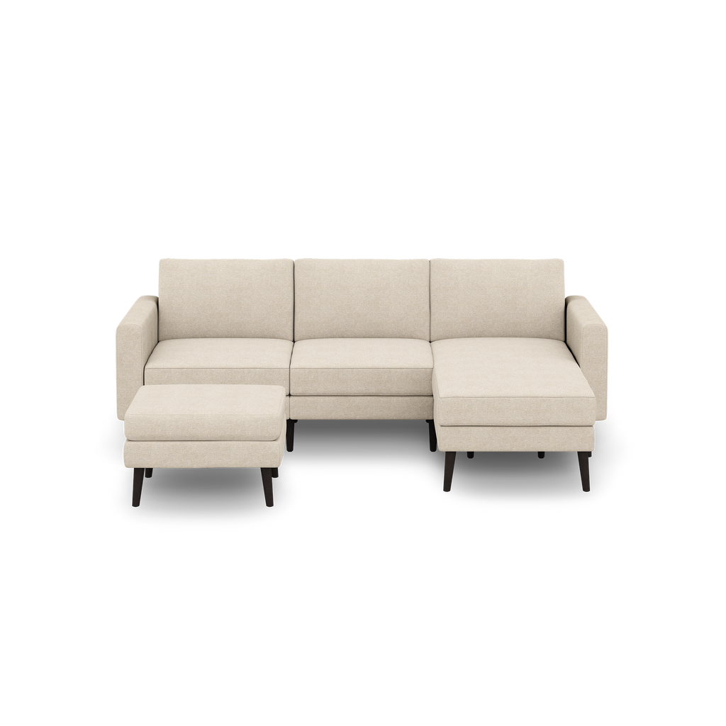 Beige fabric sectional sofa with three cushioned seats, chaise on the right side, square arms, and matching ottoman on dark wooden legs.