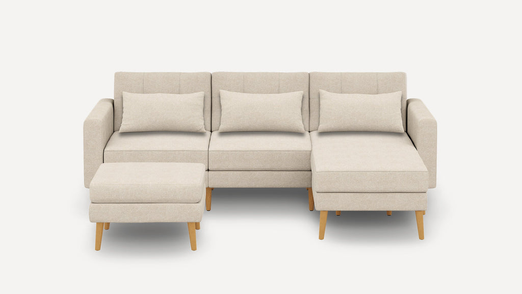 Beige fabric sectional sofa with three back cushions, wooden legs, and a matching ottoman on a white background.